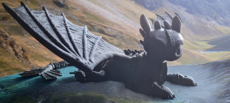 K&ouml;nnte beinhalten: Ein schwarzes 3D-gedrucktes Modell eines Drachen mit ausgebreiteten Fl&uuml;geln. Der Drache liegt auf einer gr&uuml;nen Oberfl&auml;che mit einem Berg im Hintergrund.