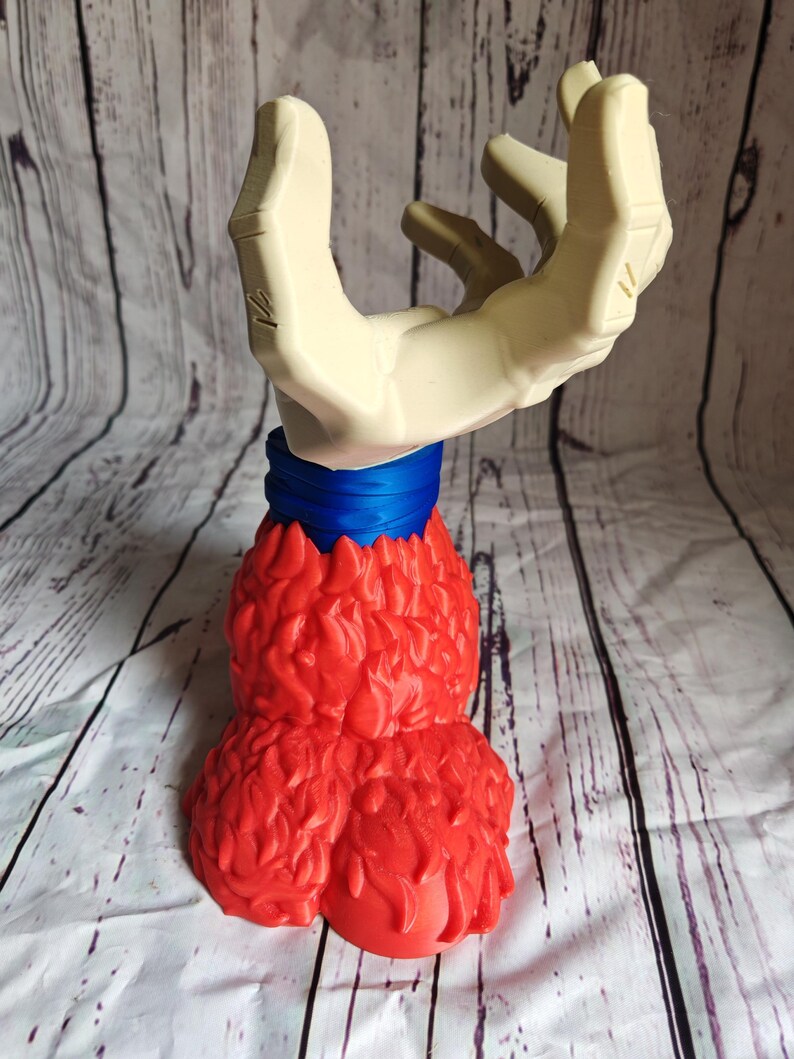 Pode incluir: Uma escultura &uacute;nica com uma m&atilde;o aberta estilizada em cor neutra, emergindo de uma base vermelha texturizada. Uma faixa azul envolve a base da m&atilde;o. A escultura est&aacute; sobre um fundo claro com textura de madeira.