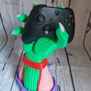 Op de afbeelding: Een groen en paars 3D-geprint beeldje van een hand die een zwarte Xbox-controller vasthoudt. De hand draagt ​​een rood en roze riem.