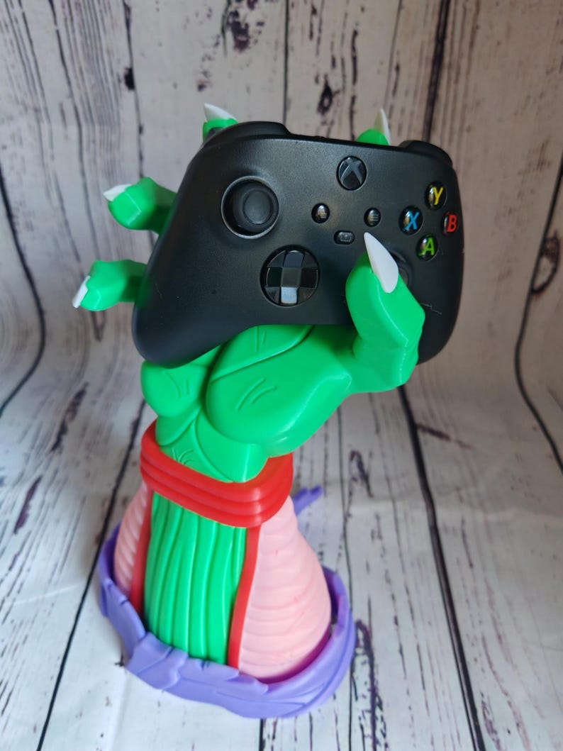 Op de afbeelding: Een groen en paars 3D-geprint beeldje van een hand die een zwarte Xbox-controller vasthoudt. De hand draagt ​​een rood en roze riem.
