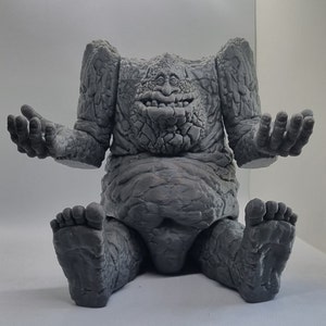 May include: A gray, stone-like figure with a large, round belly and a wide, toothy grin. The figure has two arms and two legs, and its hands are outstretched. The figure appears to be made of rough, textured stone.