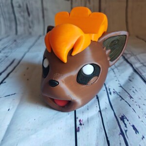 Pode incluir: Um vaso de planta de cor marrom e laranja impresso em 3D com a forma de um personagem Pokemon com uma grande crista laranja. O vaso tem uma boca aberta e olhos brancos.