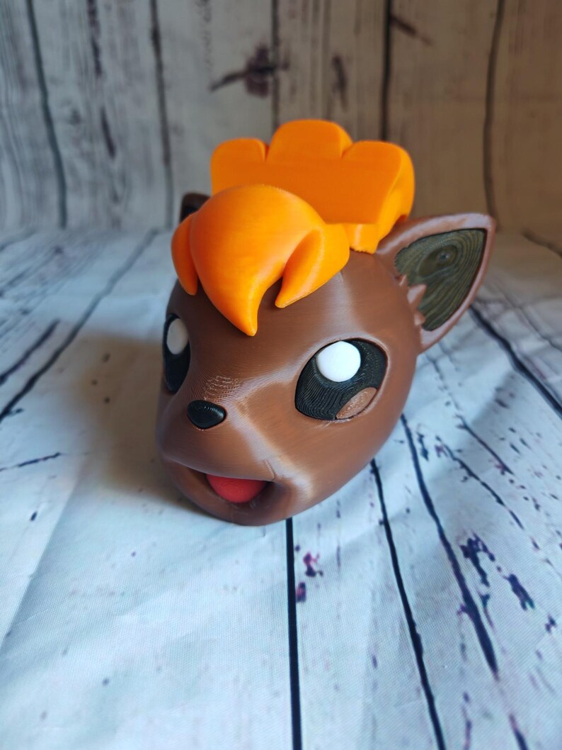 Pode incluir: Um vaso de planta de cor marrom e laranja impresso em 3D com a forma de um personagem Pokemon com uma grande crista laranja. O vaso tem uma boca aberta e olhos brancos.