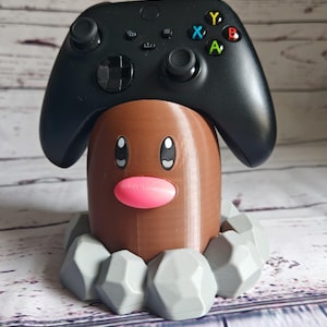 Op de afbeelding: Een 3D-geprint bruin en grijs Pok&eacute;mon-personage, dat lijkt op een Treecko, houdt een zwarte Xbox-controller vast.