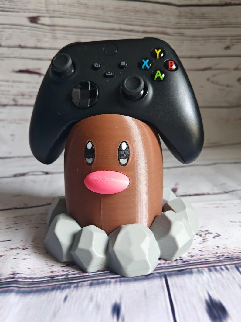Op de afbeelding: Een 3D-geprint bruin en grijs Pok&eacute;mon-personage, dat lijkt op een Treecko, houdt een zwarte Xbox-controller vast.