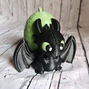 K&ouml;nnte beinhalten: Eine schwarze Drachenfigur mit gro&szlig;en Fl&uuml;geln und gr&uuml;nen Augen, vor einem hellgr&uuml;nen eif&ouml;rmigen Beh&auml;lter. Der Drache hat eine detaillierte, strukturierte Oberfl&auml;che, und das Ei hat einen kleinen Griff oben. Die Figur steht vor einem wei&szlig;-grauen Holzhintergrund.