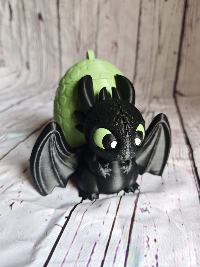 K&ouml;nnte beinhalten: Eine schwarze Drachenfigur mit gro&szlig;en Fl&uuml;geln und gr&uuml;nen Augen, vor einem hellgr&uuml;nen eif&ouml;rmigen Beh&auml;lter. Der Drache hat eine detaillierte, strukturierte Oberfl&auml;che, und das Ei hat einen kleinen Griff oben. Die Figur steht vor einem wei&szlig;-grauen Holzhintergrund.