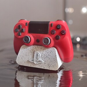 Op de afbeelding: Een rode PlayStation 4-controller staat op een witte en grijze PlayStation-thema oplaadstandaard. De standaard heeft het PlayStation-logo aan de voorkant.