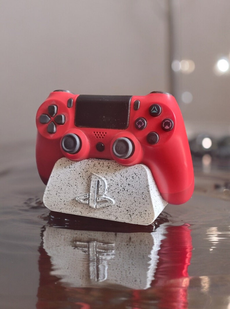 May include: A red PlayStation 4 controller is sitting on a white and gray PlayStation themed charging stand. The stand has the PlayStation logo on the front.