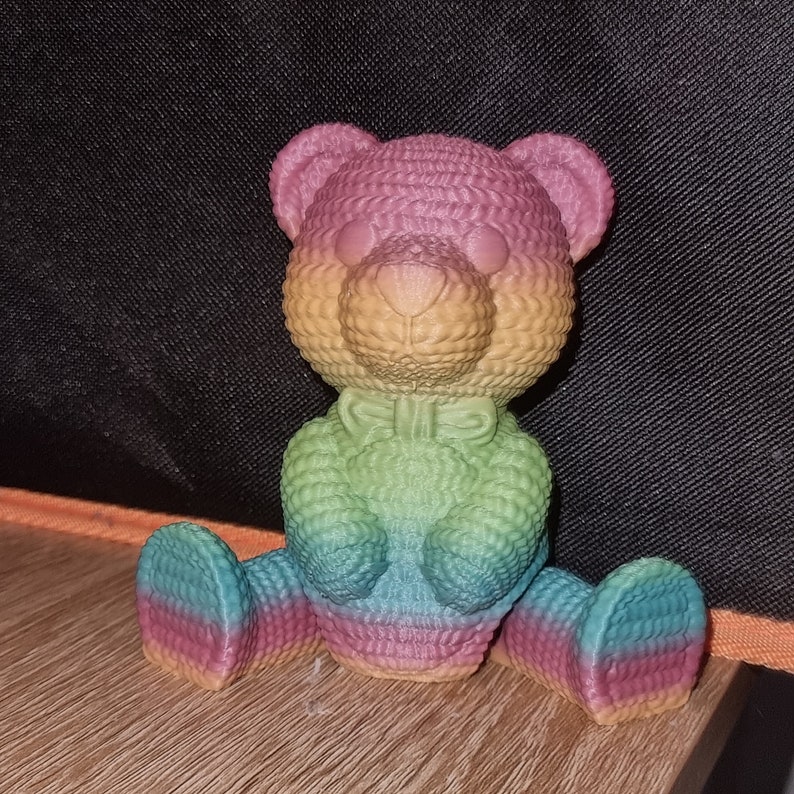 May include: A rainbow-coloured crocheted teddy bear with a bow tie. The bear is sitting on a wooden surface.