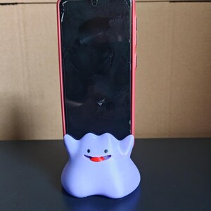 以下が含まれることがあります： 幽霊タイプのポケモンの形をした、紫色の3Dプリントされた携帯電話スタンド。スタンドは、黒い画面の赤い携帯電話ケースを持っています。