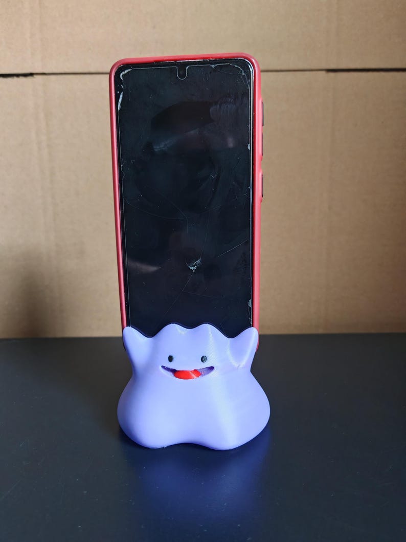 以下が含まれることがあります： 幽霊タイプのポケモンの形をした、紫色の3Dプリントされた携帯電話スタンド。スタンドは、黒い画面の赤い携帯電話ケースを持っています。