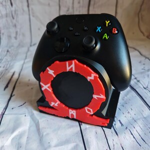 Peut inclure: Une manette Xbox noire avec des accents rouges et blancs est pos&eacute;e sur un support noir avec un logo God of War rouge et blanc. La manette poss&egrave;de les boutons Xbox standards : A, B, X, Y.