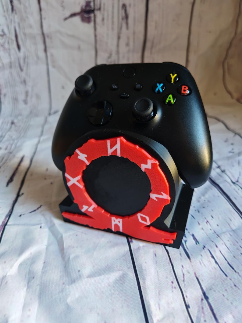 Peut inclure: Une manette Xbox noire avec des accents rouges et blancs est pos&eacute;e sur un support noir avec un logo God of War rouge et blanc. La manette poss&egrave;de les boutons Xbox standards : A, B, X, Y.
