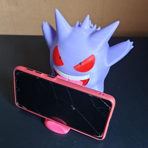 Może przedstawiać: Fioletowy uchwyt na telefon w kształcie Gengara z czerwonymi oczami i szerokim uśmiechem, trzymający pęknięty telefon w r&oacute;żowym etui. Telefon jest ustawiony poziomo. Uchwyt znajduje się na ciemnej powierzchni.