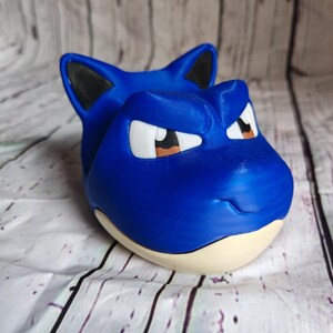 K&ouml;nnte beinhalten: Eine blaue, 3D-gedruckte Snorlax-Figur mit schwarzen Ohren, wei&szlig;en Augen und braunen Pupillen. Die Figur hat einen hellbeigen Sockel und steht vor einem wei&szlig;-grauen Holzmaserungshintergrund. Die Figur ist ein Sammlerst&uuml;ck.