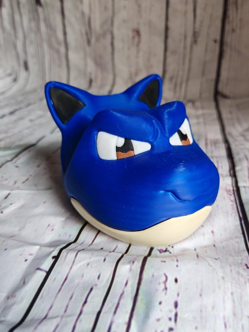 K&ouml;nnte beinhalten: Eine blaue, 3D-gedruckte Snorlax-Figur mit schwarzen Ohren, wei&szlig;en Augen und braunen Pupillen. Die Figur hat einen hellbeigen Sockel und steht vor einem wei&szlig;-grauen Holzmaserungshintergrund. Die Figur ist ein Sammlerst&uuml;ck.
