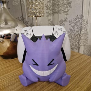 May include: A purple and white 3D printed stand for a video game controller. The stand is shaped like a Pokemon character with a wide grin.