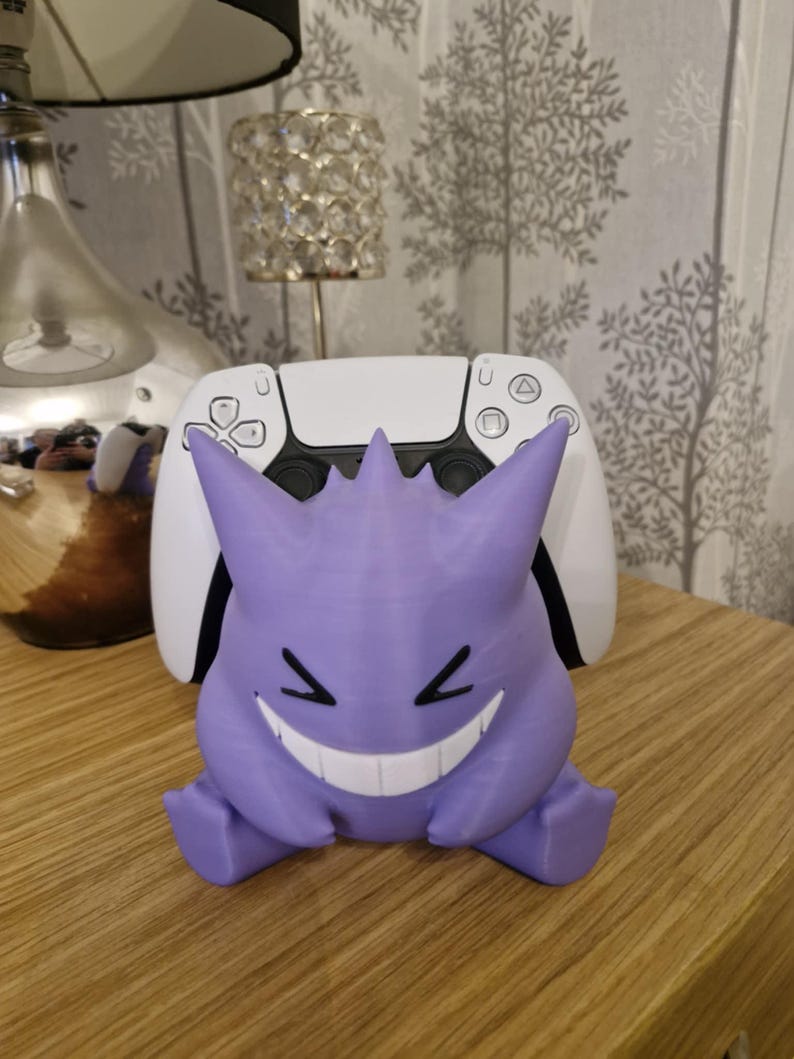 May include: A purple and white 3D printed stand for a video game controller. The stand is shaped like a Pokemon character with a wide grin.