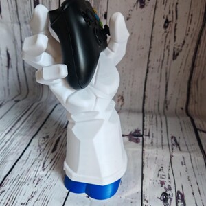 K&ouml;nnte beinhalten: Eine wei&szlig;e, im 3D-Druck hergestellte Hand h&auml;lt einen schwarzen Videospiel-Controller. Die Hand steht auf einem blauen Sockel.