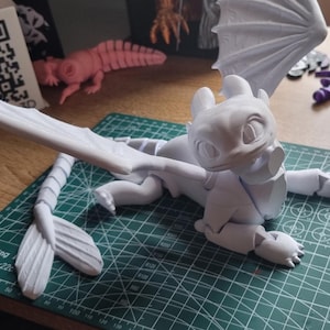 May include: A white articulated dragon figurine with wings, tail, and legs. The dragon is posed in a lying down position with its head turned to the side. The figurine is made of plastic and has a smooth, matte finish.