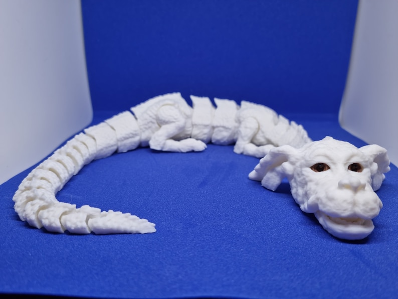 Op de afbeelding: Witte gearticuleerde drakenfiguur met een gesegmenteerd lichaam en een gedetailleerd hoofd. De draak ligt op een blauwe ondergrond.