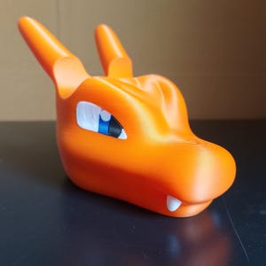 Op de afbeelding: Oranje plastic figuur van een drakenkop met een blauw en wit oog. De draak heeft een tandenrij.