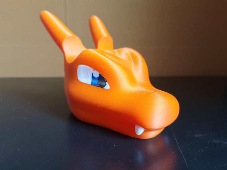 Op de afbeelding: Oranje plastic figuur van een drakenkop met een blauw en wit oog. De draak heeft een tandenrij.