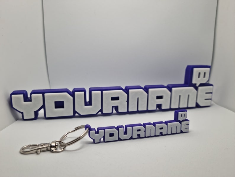 May include: A purple and white keychain with the text "YOURNAME" and a Twitch logo. The keychain is attached to a silver key ring.