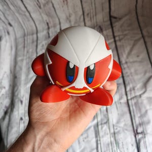 Pode incluir: Uma figura vermelha e branca de um personagem com olhos grandes e um design semelhante a uma m&aacute;scara. A figura tem um corpo redondo, bra&ccedil;os e p&eacute;s pequenos. A figura &eacute; segurada em uma m&atilde;o.