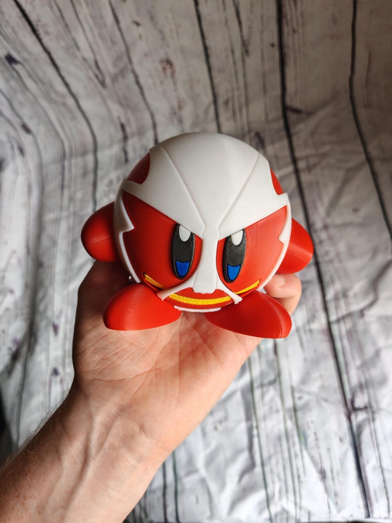 Pode incluir: Uma figura vermelha e branca de um personagem com olhos grandes e um design semelhante a uma m&aacute;scara. A figura tem um corpo redondo, bra&ccedil;os e p&eacute;s pequenos. A figura &eacute; segurada em uma m&atilde;o.