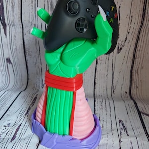 Può includere: Una statuetta stampata in 3D verde e rosa di una mano che tiene un controller Xbox nero. La mano indossa una fascia rosso e verde e ha unghie lunghe e bianche.