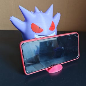 Może przedstawiać: Fioletowy uchwyt na telefon w kształcie Gengara z czerwonymi oczami i ustami, trzymający smartfon w czerwonej ramce. Telefon jest pęknięty. Uchwyt stoi na czarnej powierzchni.