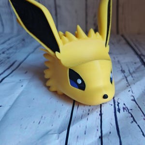 Peut inclure: Une figurine en 3D imprim&eacute;e en jaune d'un personnage de Pokemon avec des oreilles noires et des yeux bleus. La figurine est sur une surface en bois blanc.