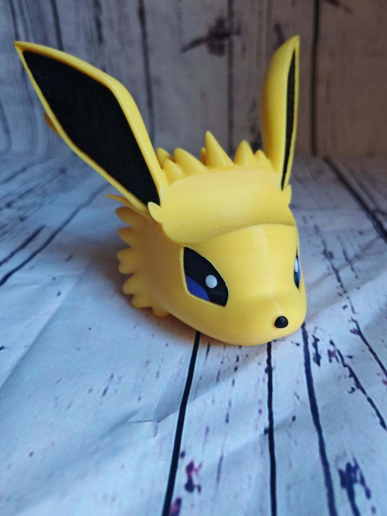 Peut inclure: Une figurine en 3D imprim&eacute;e en jaune d'un personnage de Pokemon avec des oreilles noires et des yeux bleus. La figurine est sur une surface en bois blanc.