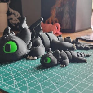 May include: Two black dragon figurines with green eyes. The larger dragon is lying down with its head turned to the side. The smaller dragon is lying down with its head facing forward. Both dragons are made of a smooth, black material.