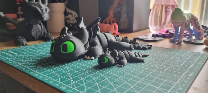 May include: Two black dragon figurines with green eyes. The larger dragon is lying down with its head turned to the side. The smaller dragon is lying down with its head facing forward. Both dragons are made of a smooth, black material.