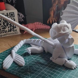 K&ouml;nnte beinhalten: Wei&szlig;e 3D-gedruckte Drachenfigur mit langem Schwanz, Fl&uuml;geln und einem freundlichen Gesicht. Der Drache liegt auf dem Bauch mit ausgebreiteten Fl&uuml;geln. Die Figur befindet sich auf einer gr&uuml;nen Schneidematte.