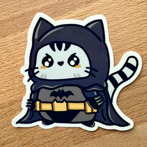 May include: A die-cut sticker featuring a cartoon cat dressed as Batman. The cat is wearing a dark gray and black costume with a yellow utility belt and a bat symbol. The cat has a white face with large, cute eyes and a striped tail.