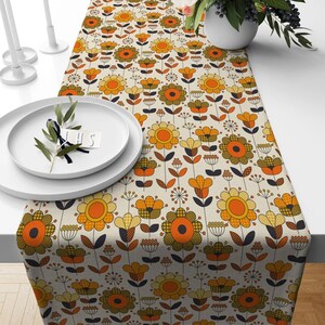May include: A floral table runner with a retro design. The runner features a repeating pattern of orange, yellow, and brown flowers on a cream background. The flowers have a geometric style, with some featuring checkered patterns. The runner is displayed on a white table.