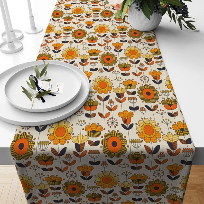 May include: A floral table runner with a retro design. The runner features a repeating pattern of orange, yellow, and brown flowers on a cream background. The flowers have a geometric style, with some featuring checkered patterns. The runner is displayed on a white table.