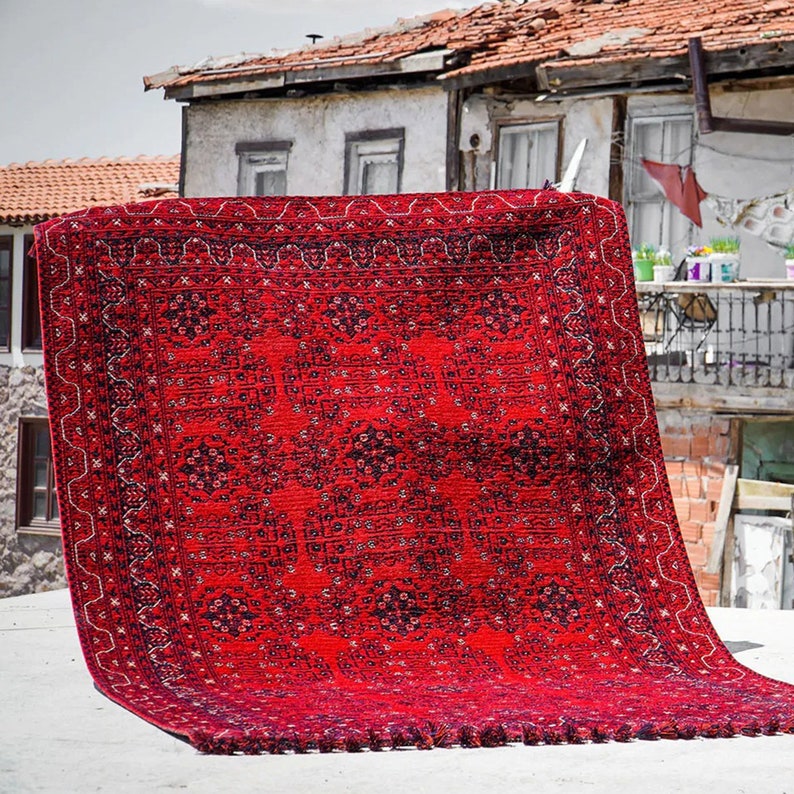Pu&ograve; includere: Un tappeto rosso con un design persiano tradizionale. Il tappeto ha un bordo blu scuro e motivi intricati su tutta la superficie. Il tappeto &egrave; steso su una superficie di cemento.