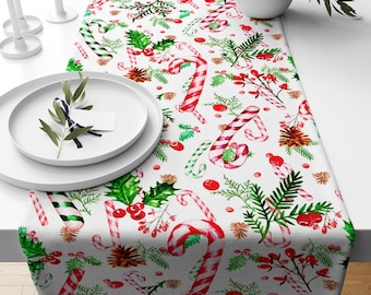 May include: A white Christmas table runner with a festive pattern of red and green candy canes, holly, pinecones, and berries. The runner is decorated with a repeating pattern of holiday elements, perfect for a seasonal dining table.
