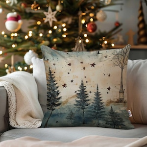 Könnte beinhalten: Quadratisches Kissen mit einer winterlichen Aquarellszene. Das Design zeigt dunkelgrüne Kiefern, einen hohen kahlen Baum und ein kleines Gebäude unter einem Sternenhimmel. Das Kissen liegt auf einem grauen Sofa mit einer weißen Decke.