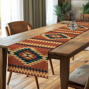 May include: A rectangular table runner with a geometric pattern in shades of rust, cream, and navy blue. The runner is placed on a wooden table, adding a decorative touch to the dining area. The design features repeating diamond and zigzag shapes.