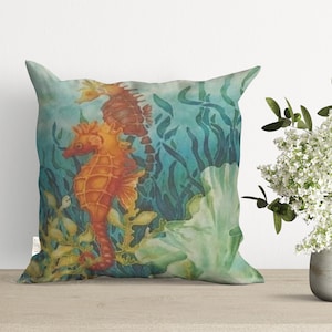 May include: A decorative pillow featuring a watercolor painting of two orange and brown seahorses swimming among green seaweed and a white coral. The background is a light blue.