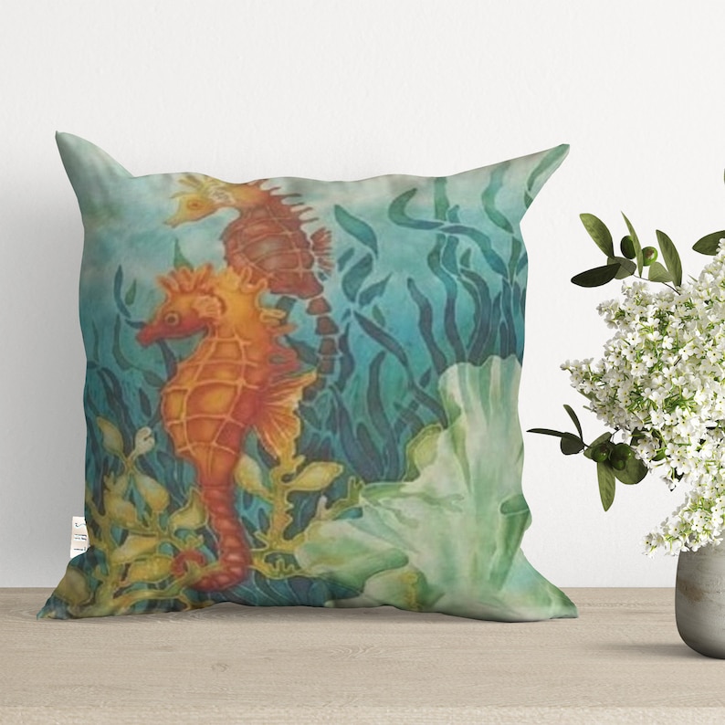 May include: A decorative pillow featuring a watercolor painting of two orange and brown seahorses swimming among green seaweed and a white coral. The background is a light blue.