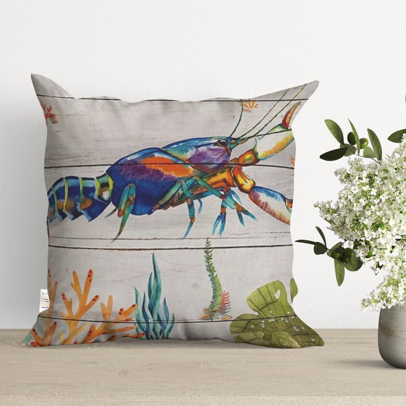 May include: A colorful pillow featuring a blue, green, orange, and purple lobster on a white wood plank background. The lobster is surrounded by coral and seaweed.