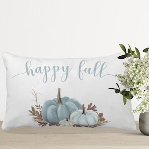 A white pillow with the text "happy fall" in blue script. The pillow features a watercolor illustration of three blue pumpkins with brown leaves and stems.