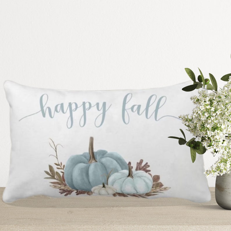 A white pillow with the text "happy fall" in blue script. The pillow features a watercolor illustration of three blue pumpkins with brown leaves and stems.